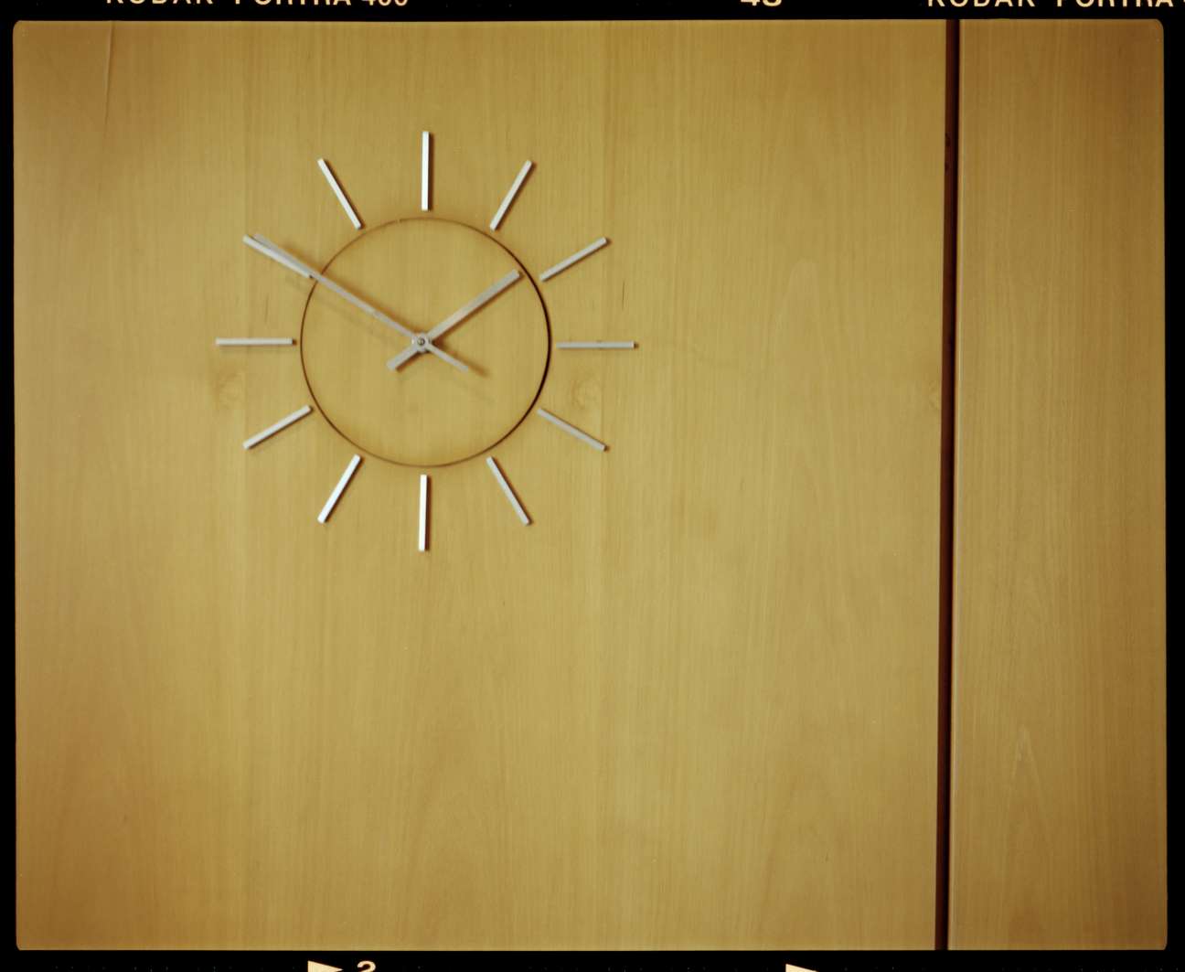 Minimalist wall-mounted clock with round brass face and chrome hour markers on wooden panel.