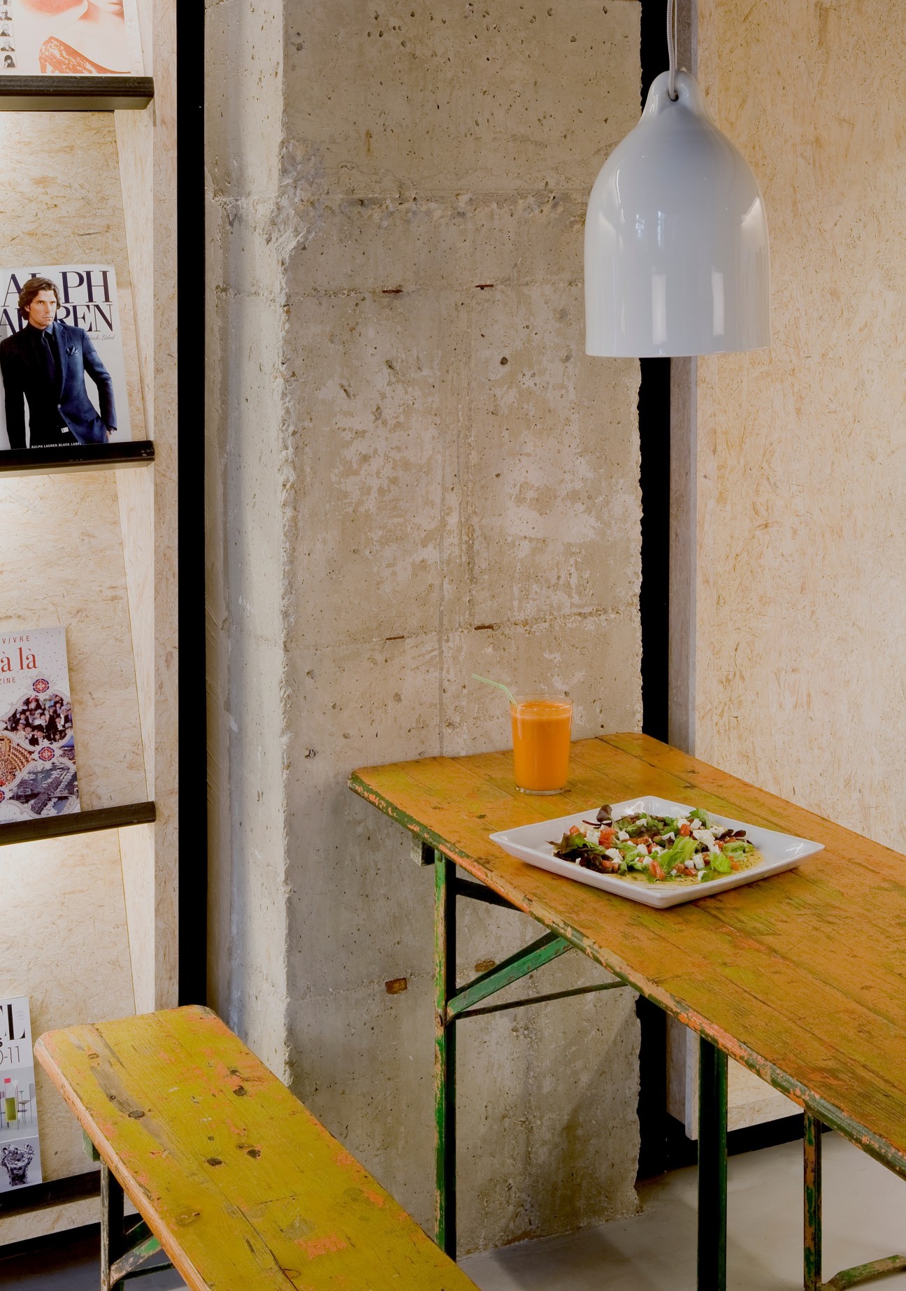 Magasand Madrid cafeteria dining area with yellow wooden table, bench seating, white pendant lamp, and concrete walls.