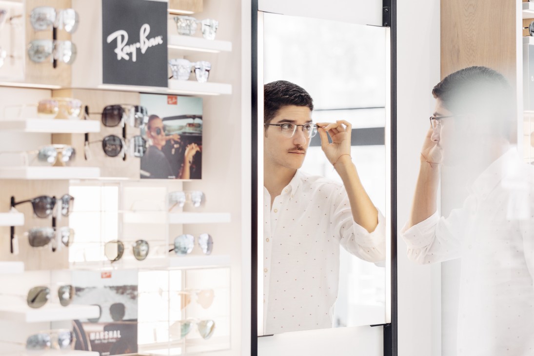 Customer trying on frameless glasses at Magrabi eyewear shop with Ray-Ban display visible.