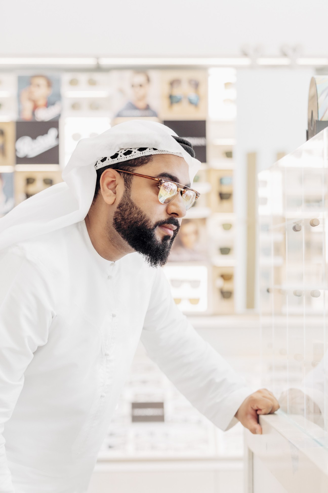 Man wearing white traditional dress and glasses examines eyewear display at Magrabi retail shop.