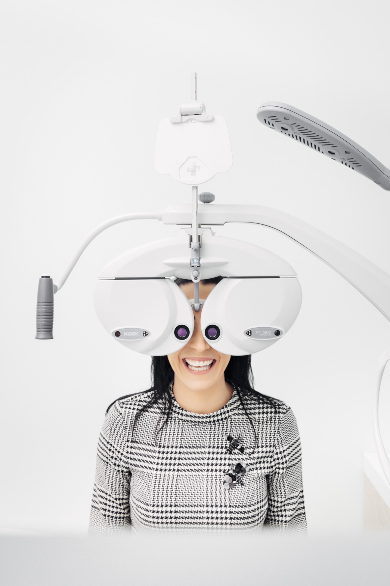 Woman wearing a houndstooth sweater smiling while undergoing an eye examination with optometry equipment.