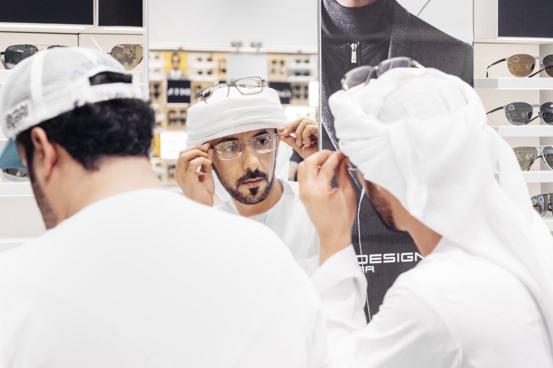 Customer trying on clear-framed glasses in a Magrabi eyewear shop with white walls and shelves of sunglasses visible in background.
