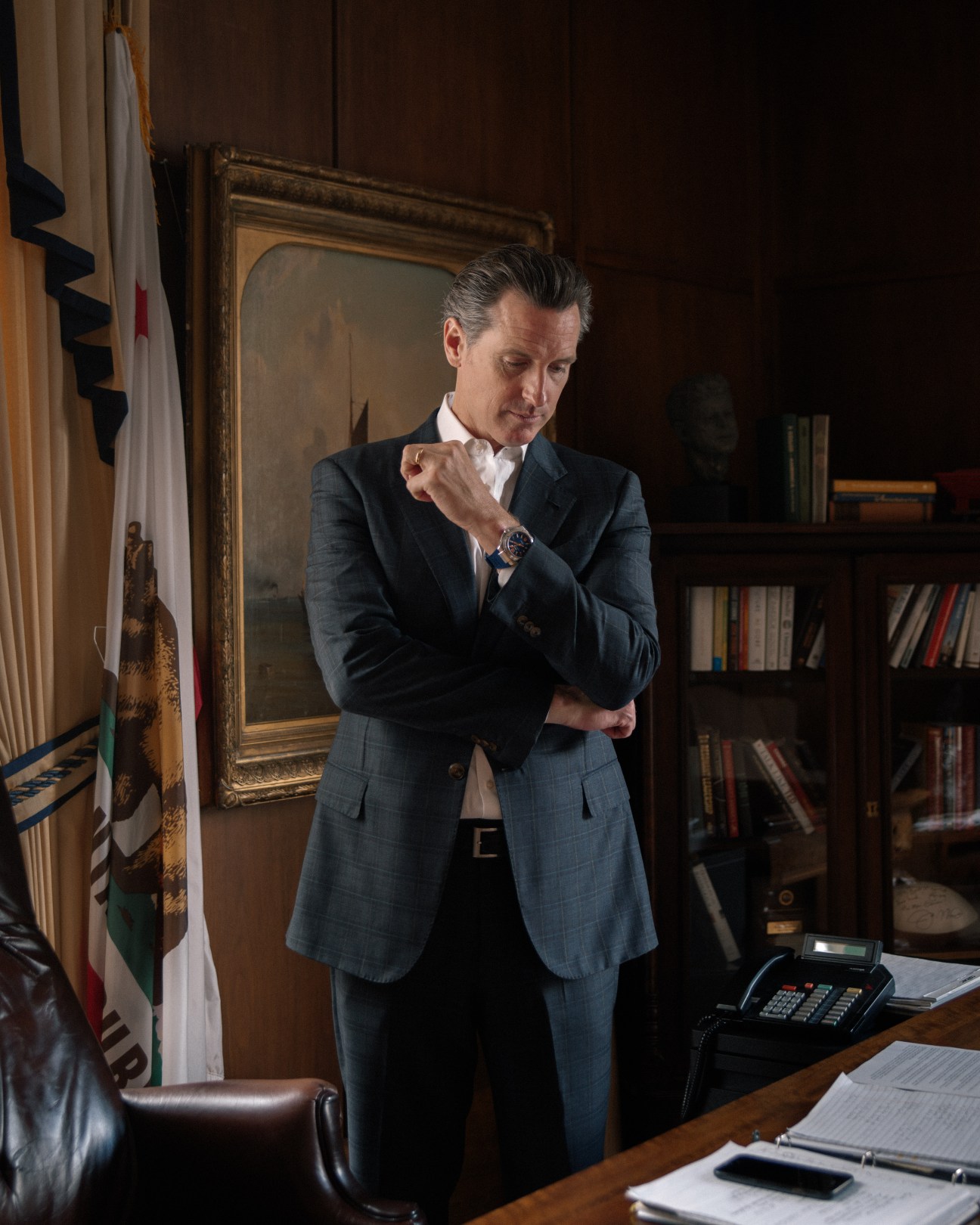 Lieutenant governor Gavin Newsom in an office with a portrait and bookshelf, wearing a blue suit and adjusting his watch.