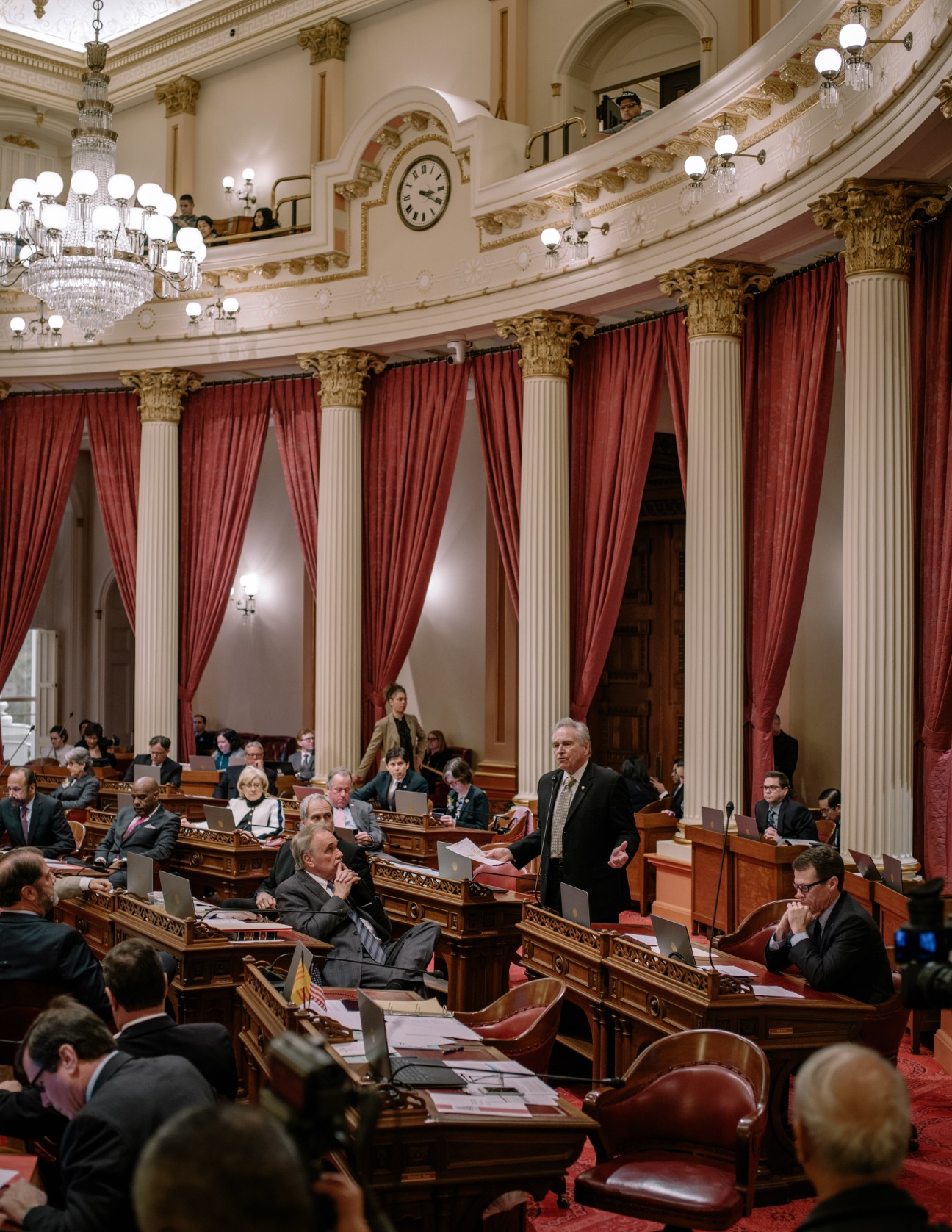 California State Senate chamber during debate with legislators seated at wooden desks, red velvet curtains, ornate gold columns and crystal chandeliers.