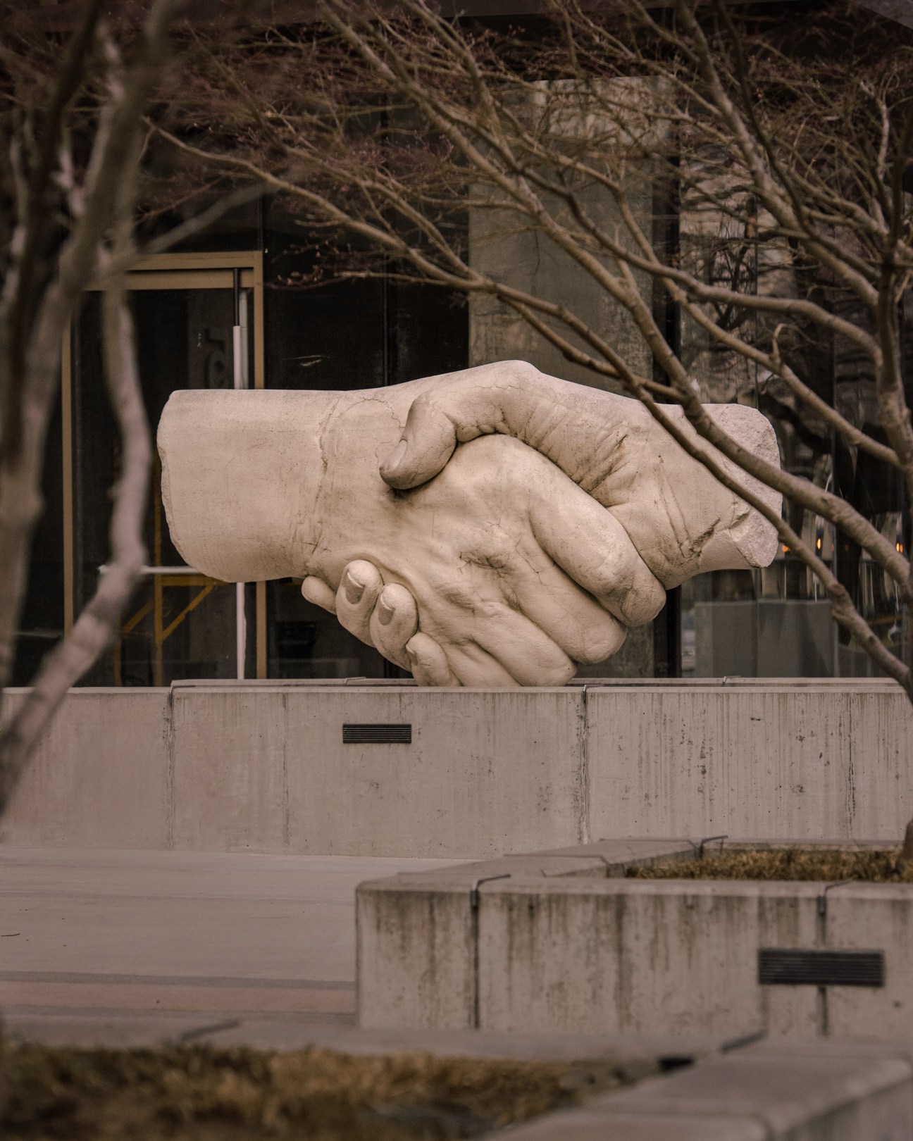 Large stone sculpture of two hands clasped in a handshake, framed by bare tree branches in an urban setting.