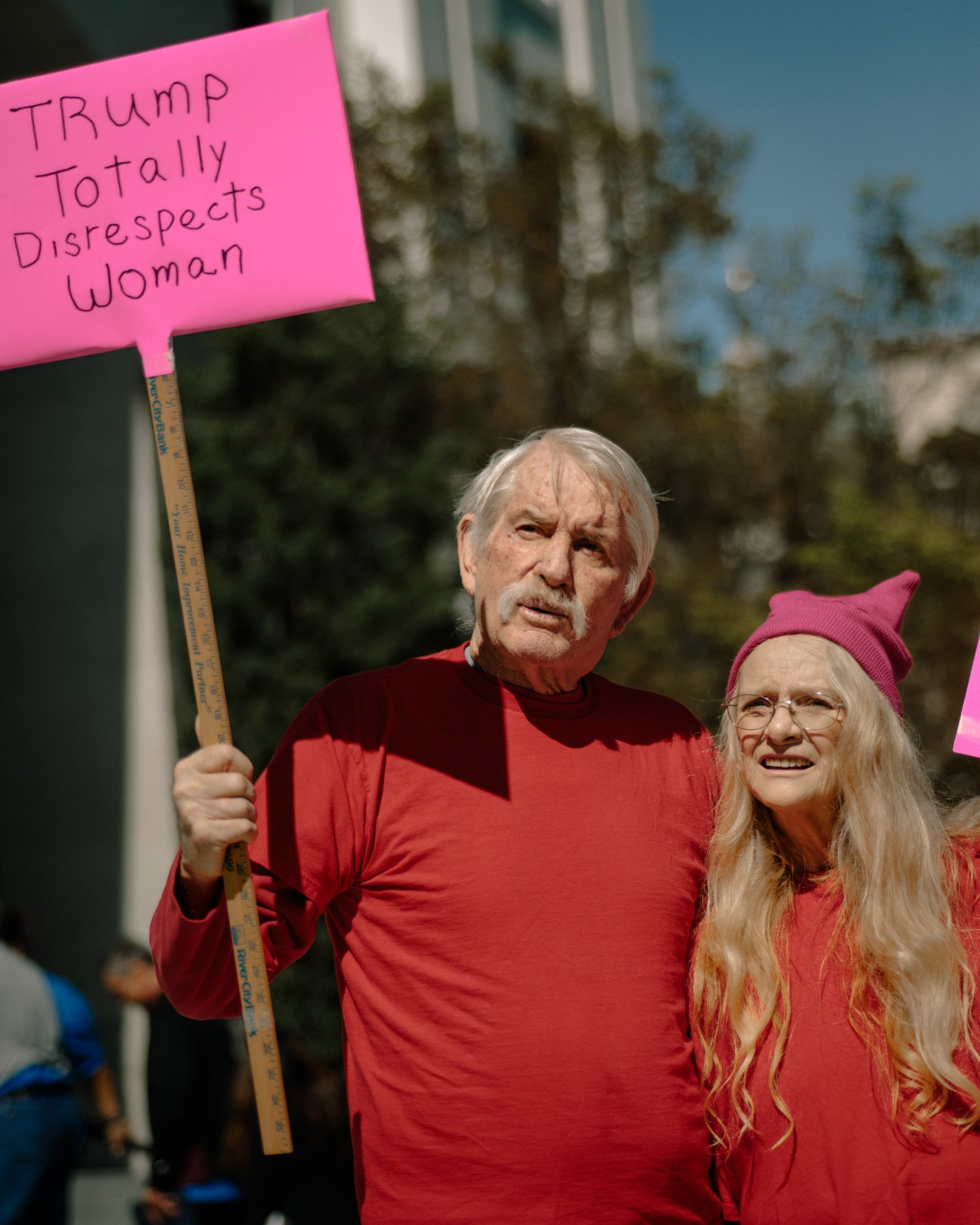 Elderly man and woman in red clothing holding a pink protest sign reading 'TRUMP Totally Disrespects Woman' at an outdoor rally.
