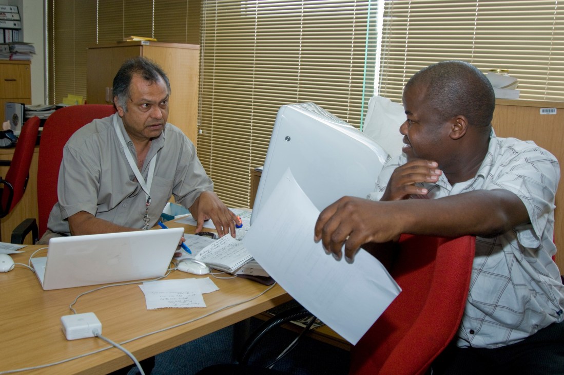 Two journalists discuss editorial content at a desk in a newspaper office.