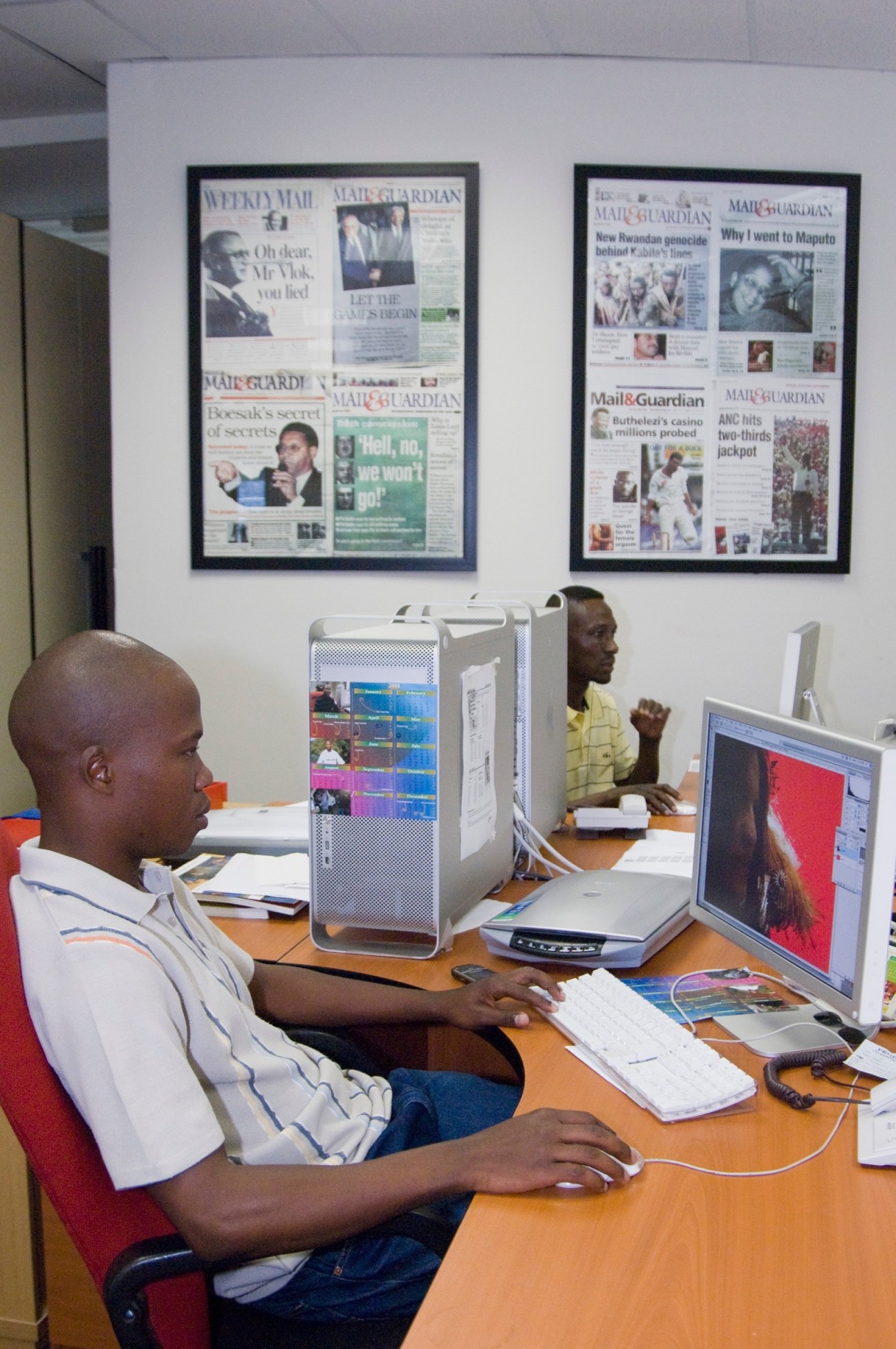 Journalists work at computers in the Mail & Guardian newsroom with framed newspaper covers displayed on the wall behind them.