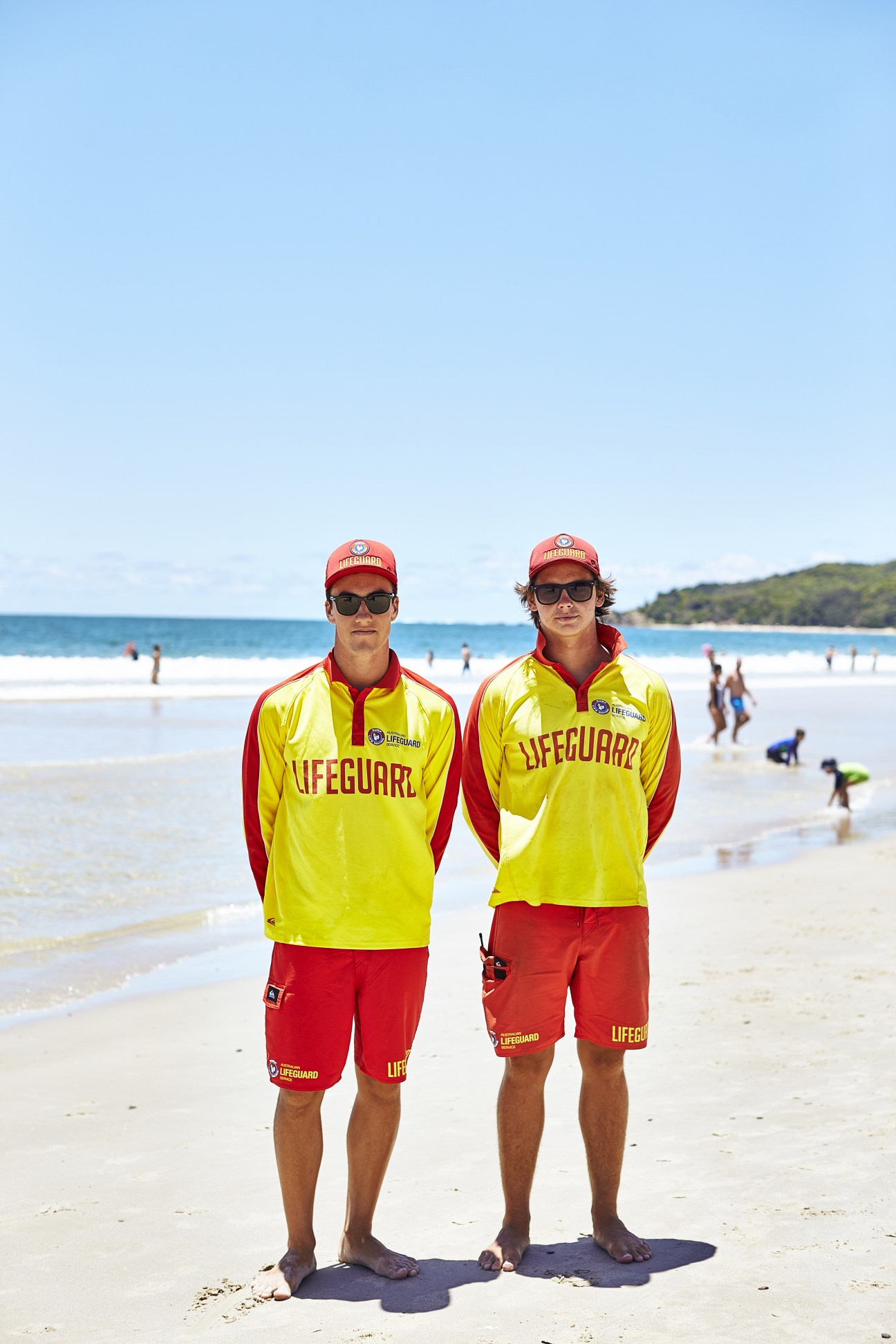 Two lifeguards in yellow and red uniforms standing on a beach with swimmers in the background.