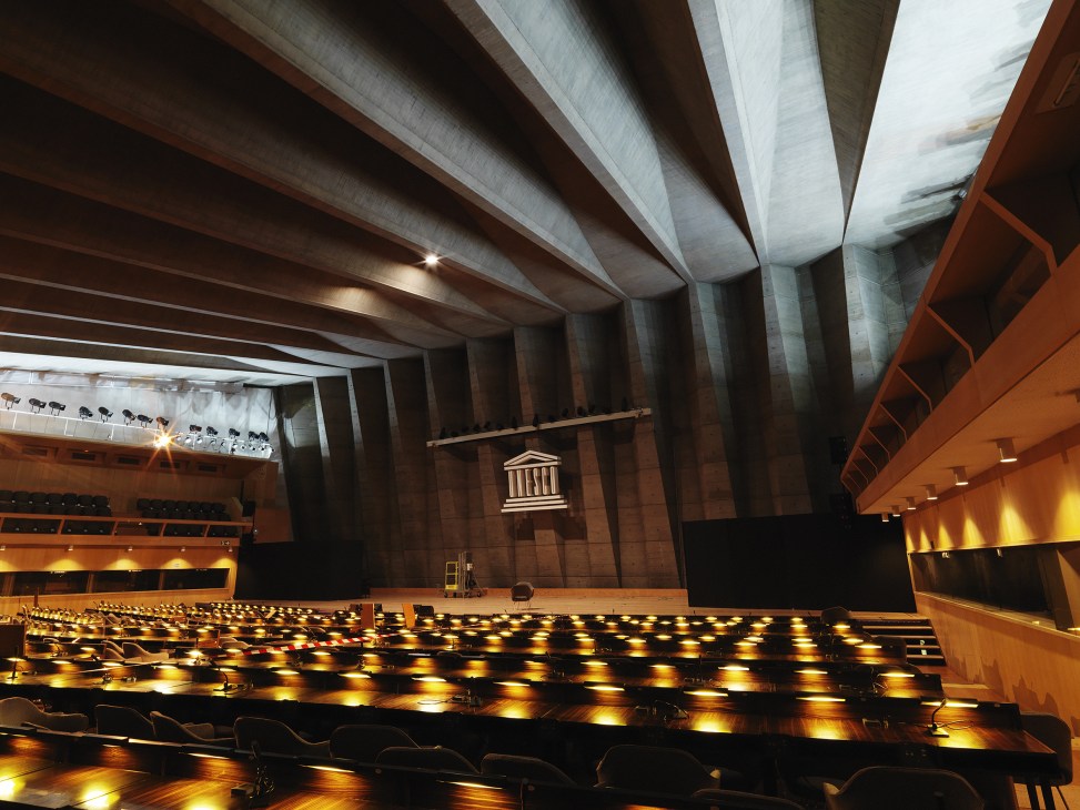 Interior of Unesco's main conference hall with rows of lit seats, brutalist concrete ceiling, and UNESCO emblem on back wall.