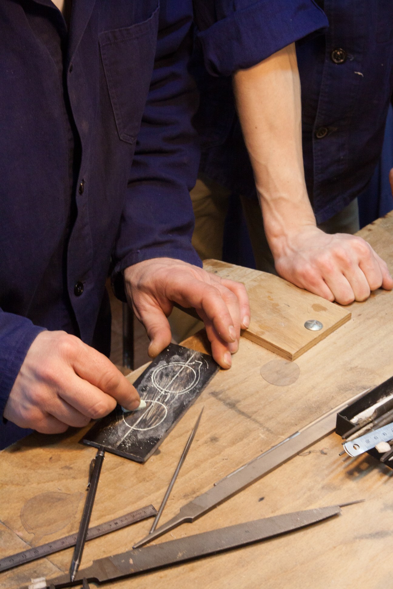 Hands tracing eyeglass frame design onto a black plastic pattern at a wooden workbench.