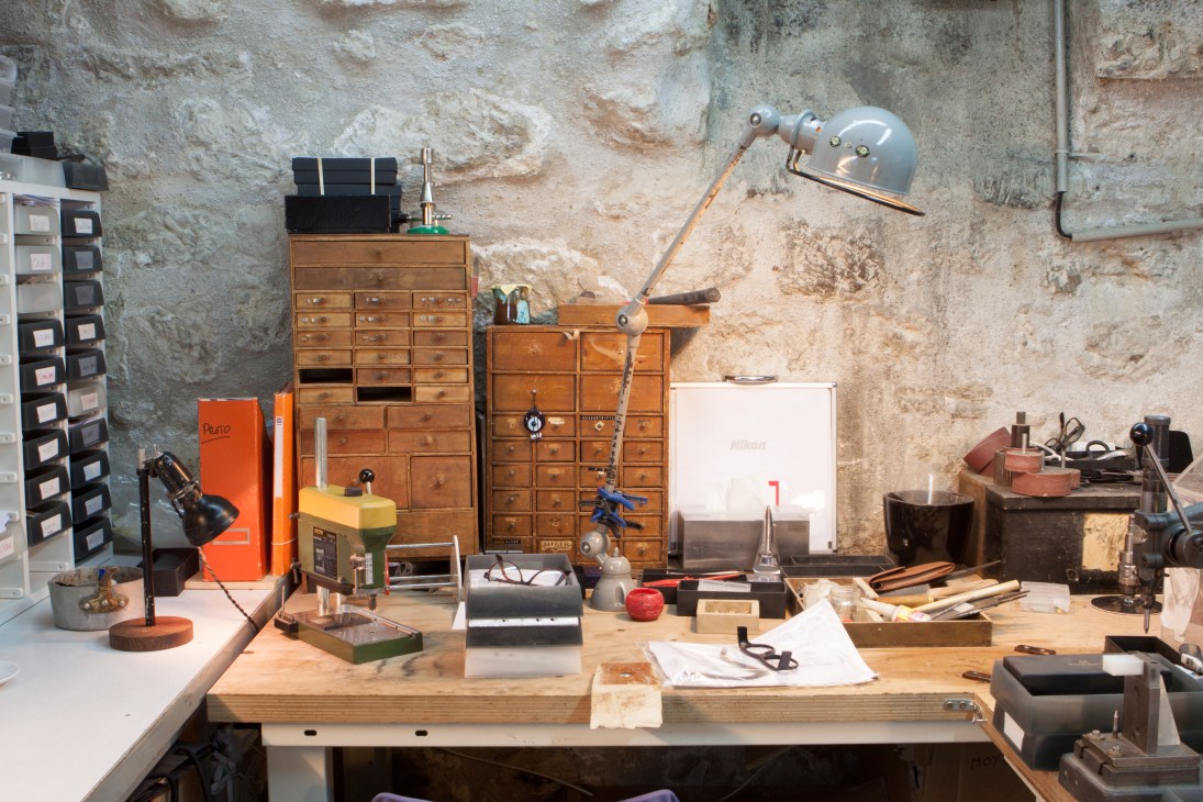 Maison Bonnet's basement atelier workspace with wooden storage drawers, craft tools, desk lamp, and materials against weathered stone walls.