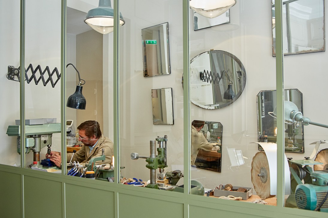 A craftsman works at a workbench in Maison Bourgeat's Parisian shop, surrounded by eyewear frames and tools.
