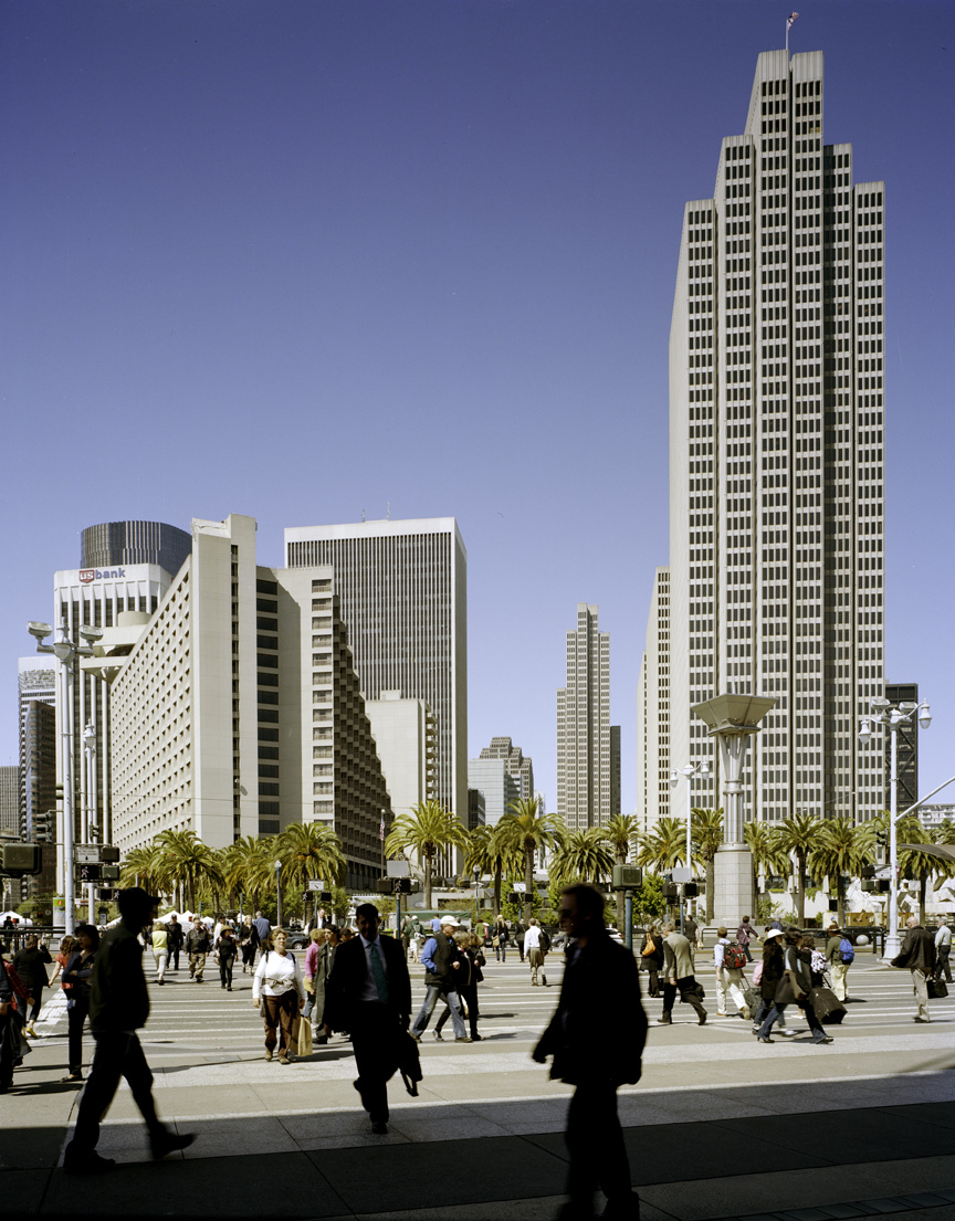 San Francisco's Embarcadero with tall downtown skyscrapers, palm trees, and pedestrians in a public plaza under clear blue sky.