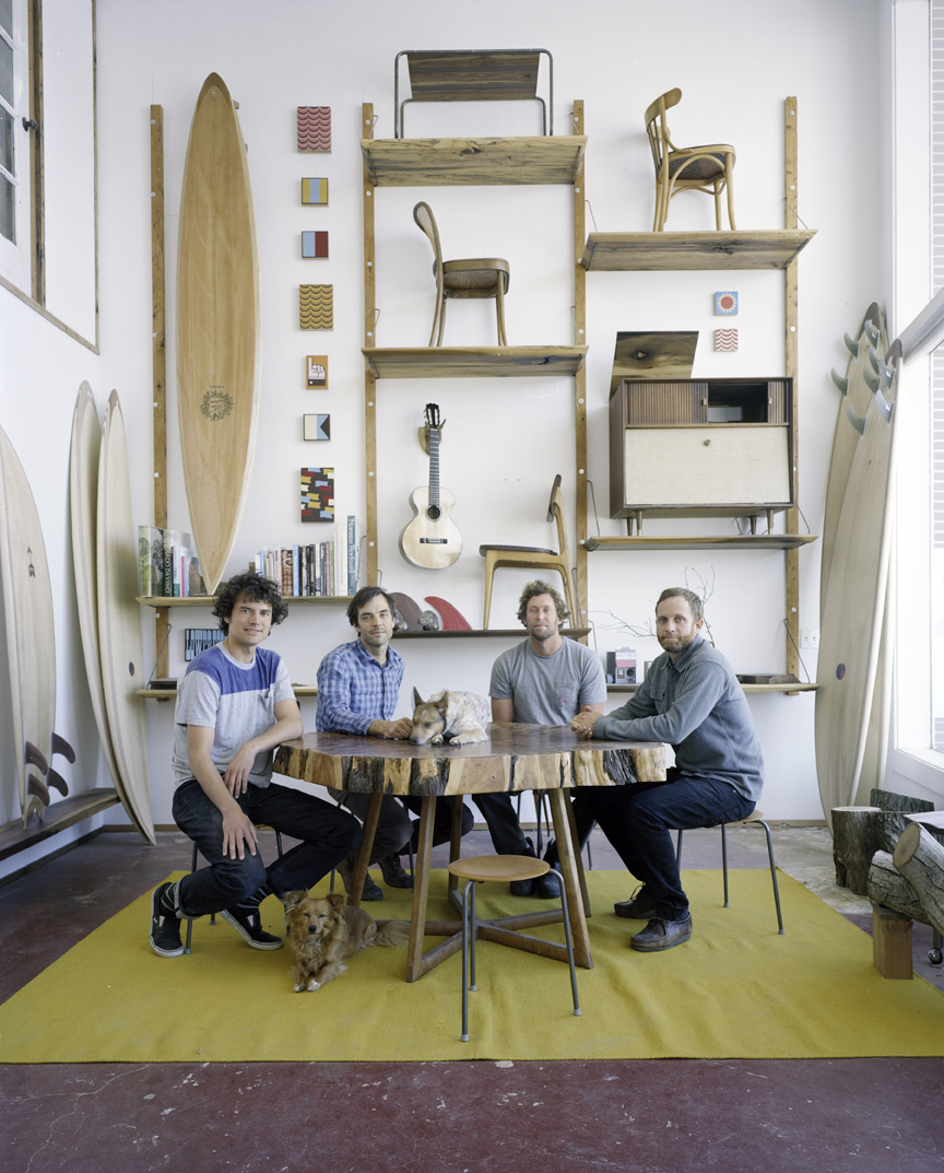Four designers pose in their workshop surrounded by wooden surfboards, furniture, and hand-painted signs displayed on shelves.