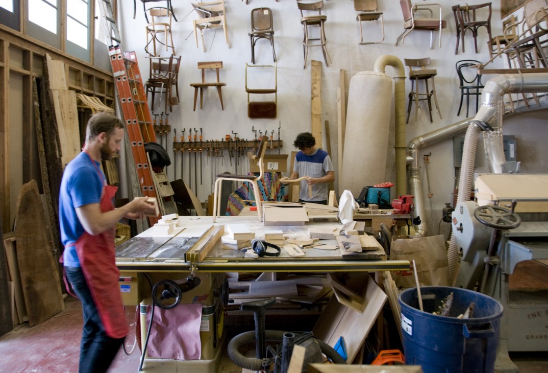 Woodworkers in a San Francisco workshop surrounded by hanging chairs, tools, and furniture projects.