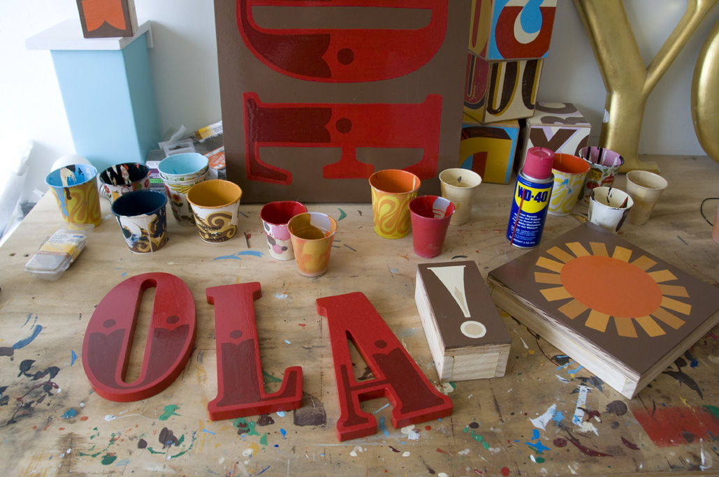 A workshop table displays red hand-painted signage letters and design materials including painted wooden pieces, brushes, and colorful cups.