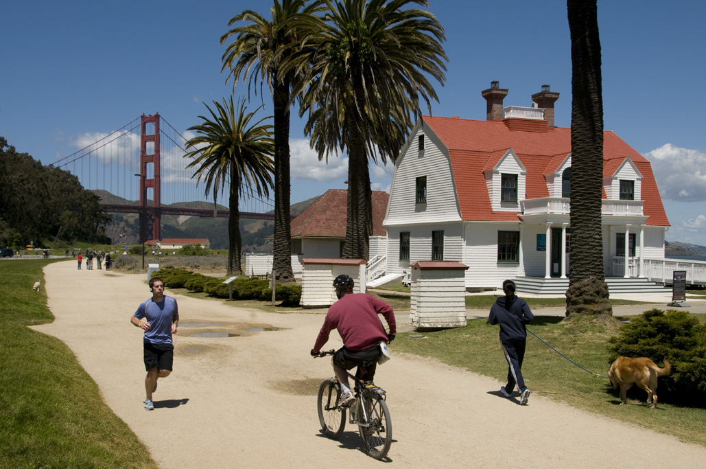 People jogging and cycling on a waterfront path with the Golden Gate Bridge and palm trees visible in the background.
