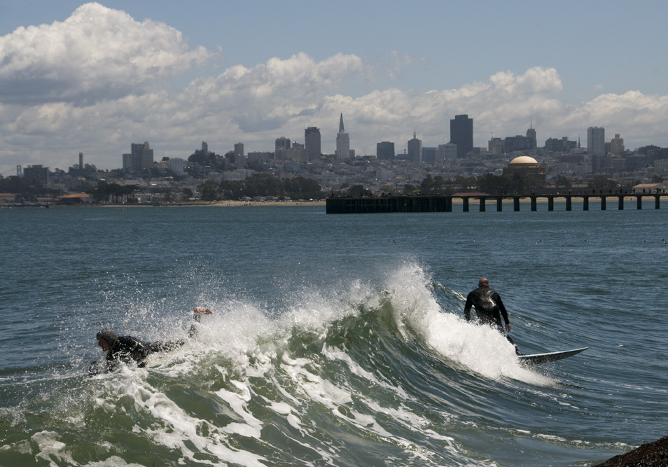 Surfers ride waves off San Francisco's coast with the city skyline and pier visible in the background.