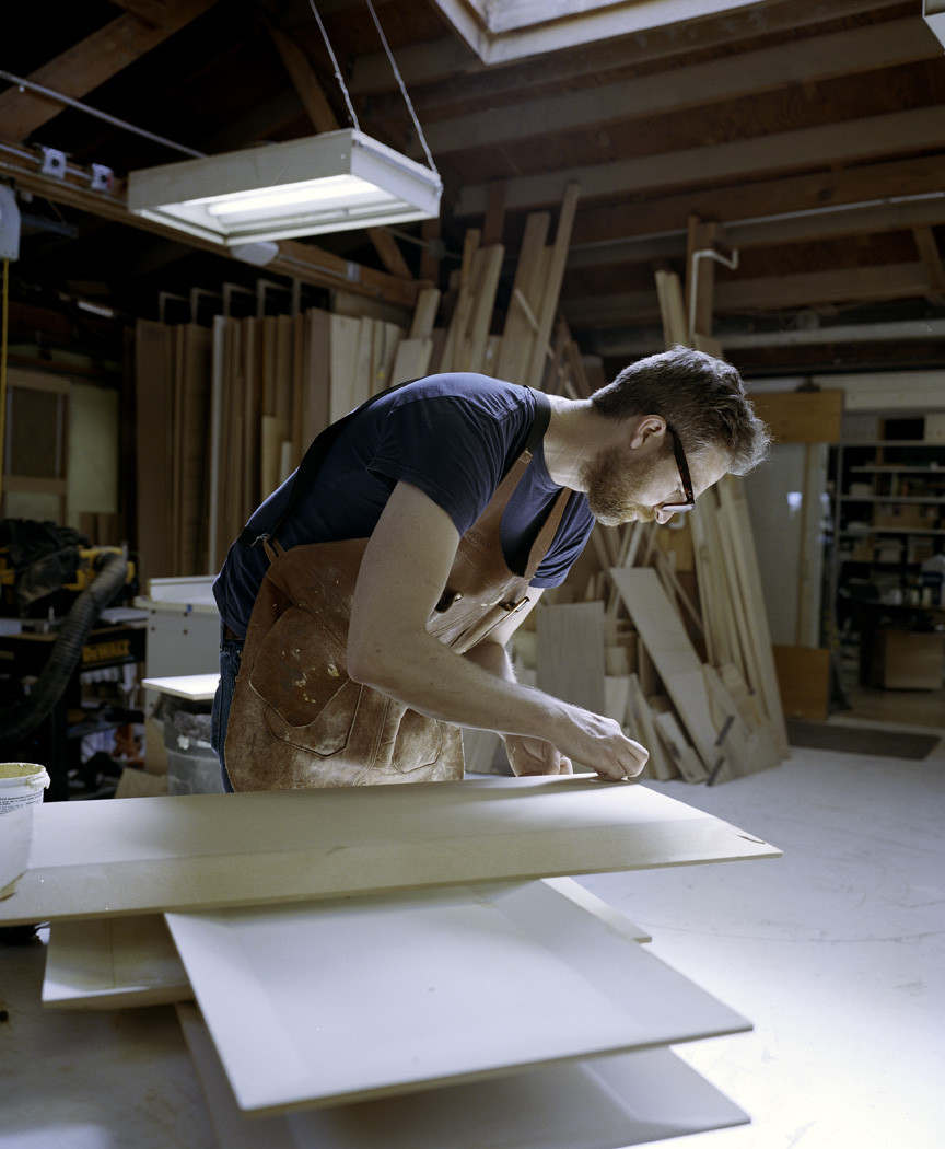 Craftsman working on white furniture pieces in a bright workshop with wooden materials and tools visible in the background.