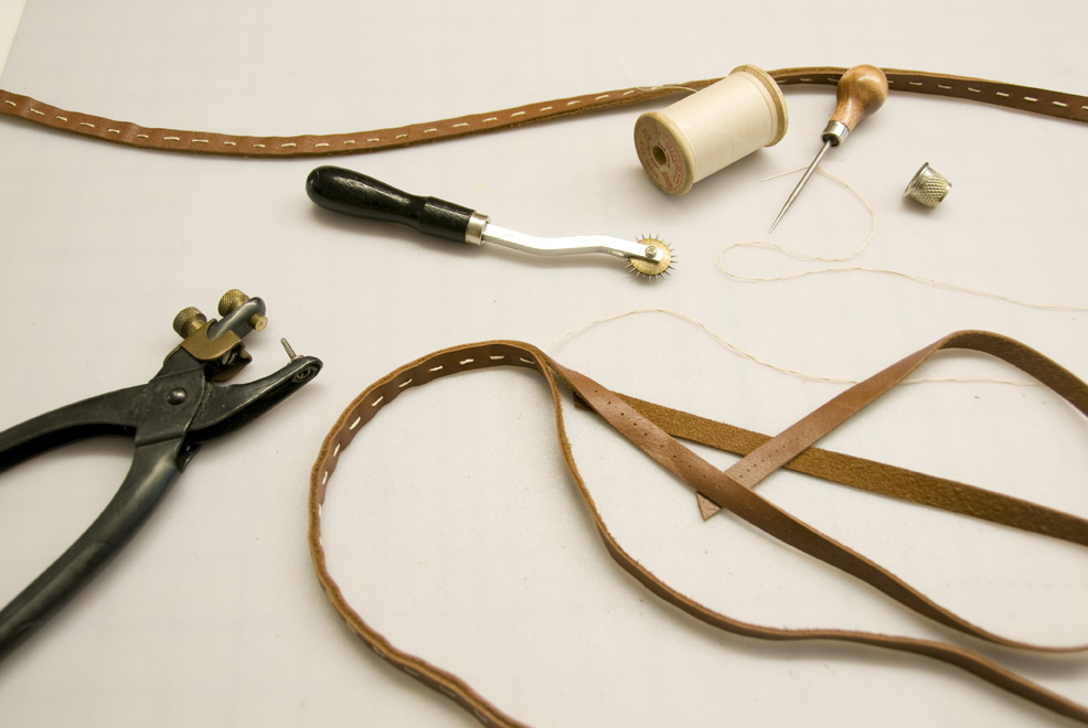 Craft tools and materials including leather straps, thread, needle, punch tool, and metal hardware on white background.