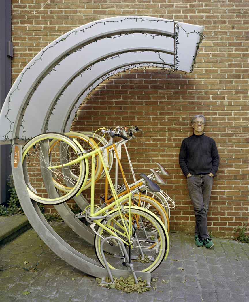 Rob Forbes stands next to yellow Public Bikes displayed against a brick wall with a modern spiral bike rack installation.