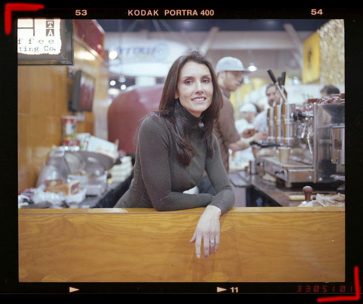 Stefania Buckley of Caffe Vita Coffee & Via Tribunali smiles at the counter of a coffee shop with espresso machines visible in the background.