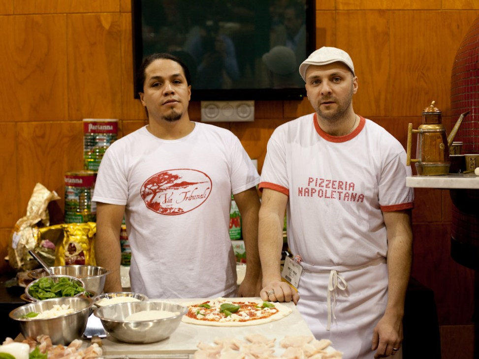 Two men in white aprons and branded t-shirts stand at a table with pizza ingredients and a finished Neapolitan pizza at the International Pizza Expo.