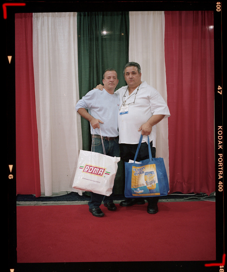 Two men in white shirts holding branded shopping bags stand against a red carpet and draped curtain backdrop.