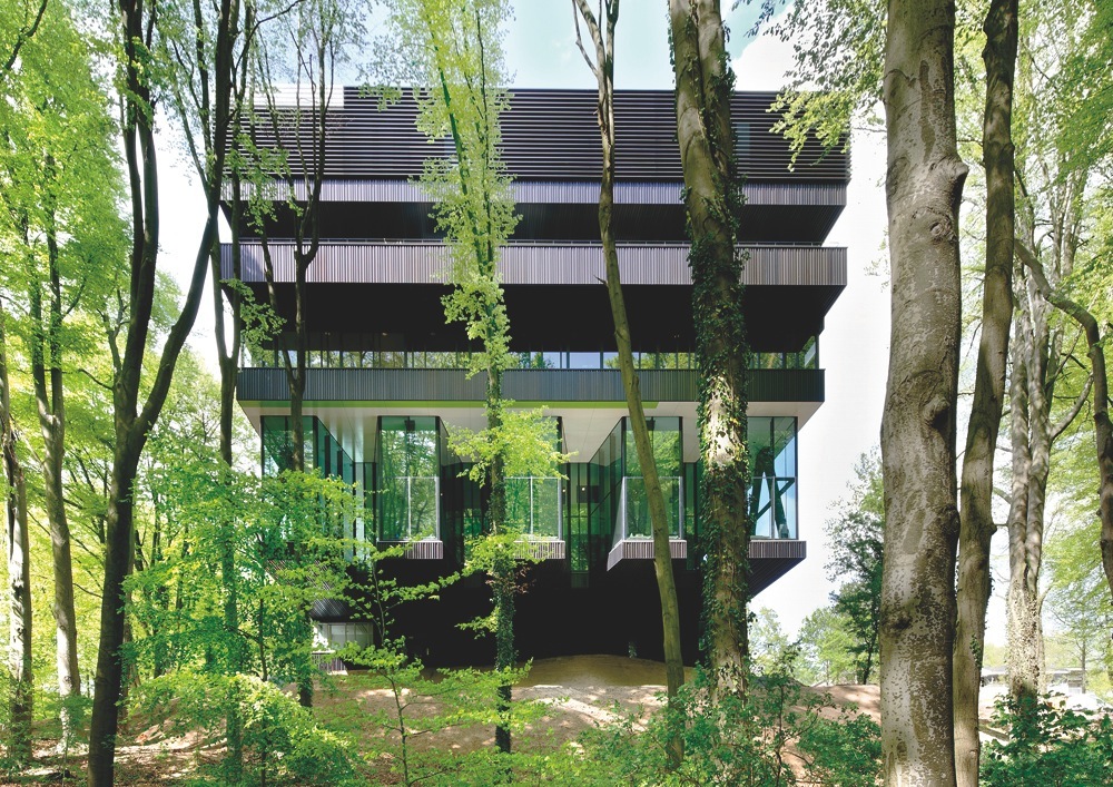 Modern multi-story rehabilitation centre nestled among tall trees with black metal framework and glass panels.