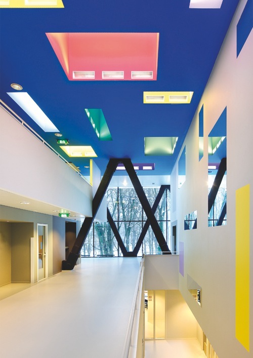 Interior hallway with colorful ceiling lights, white walls, large black X-shaped structural element, and windows overlooking trees.