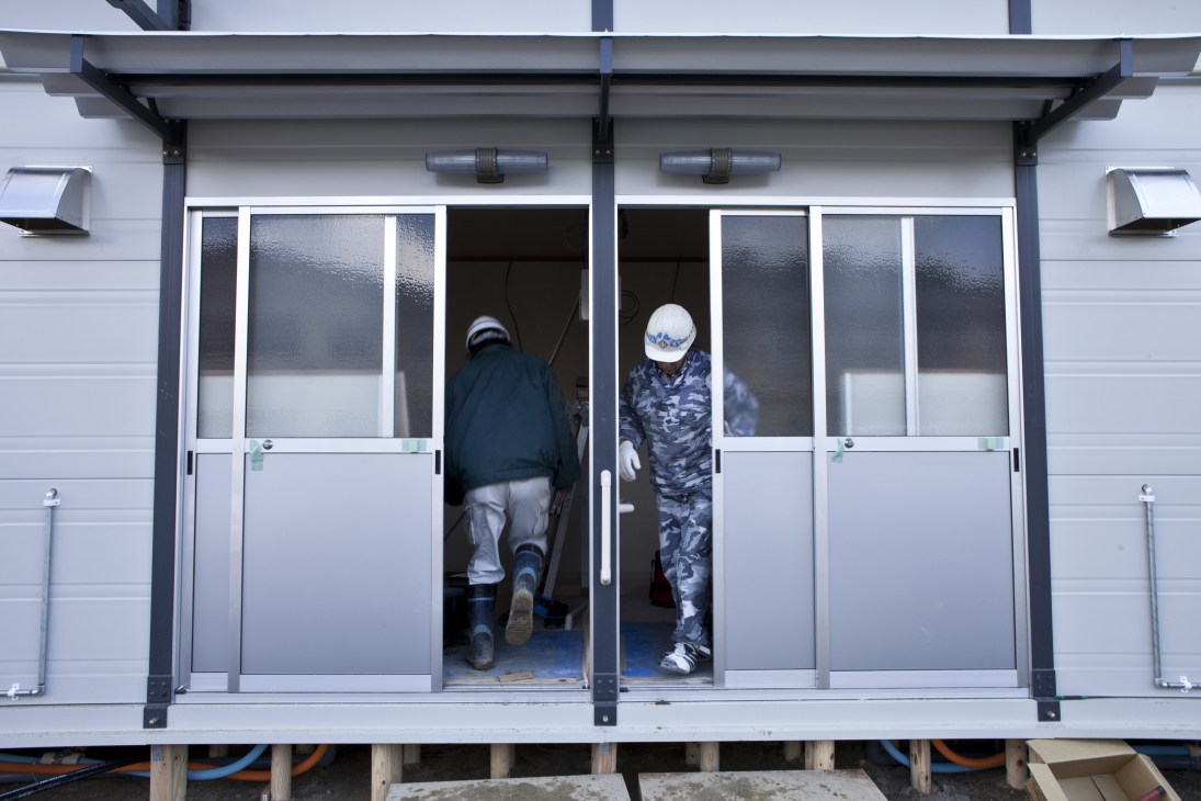 Two workers in hard hats and safety gear stand inside a prefabricated temporary housing unit with large windows and modern fixtures.