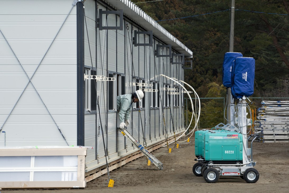 Construction worker installing utilities on a prefabricated temporary housing unit with a generator visible in the foreground.
