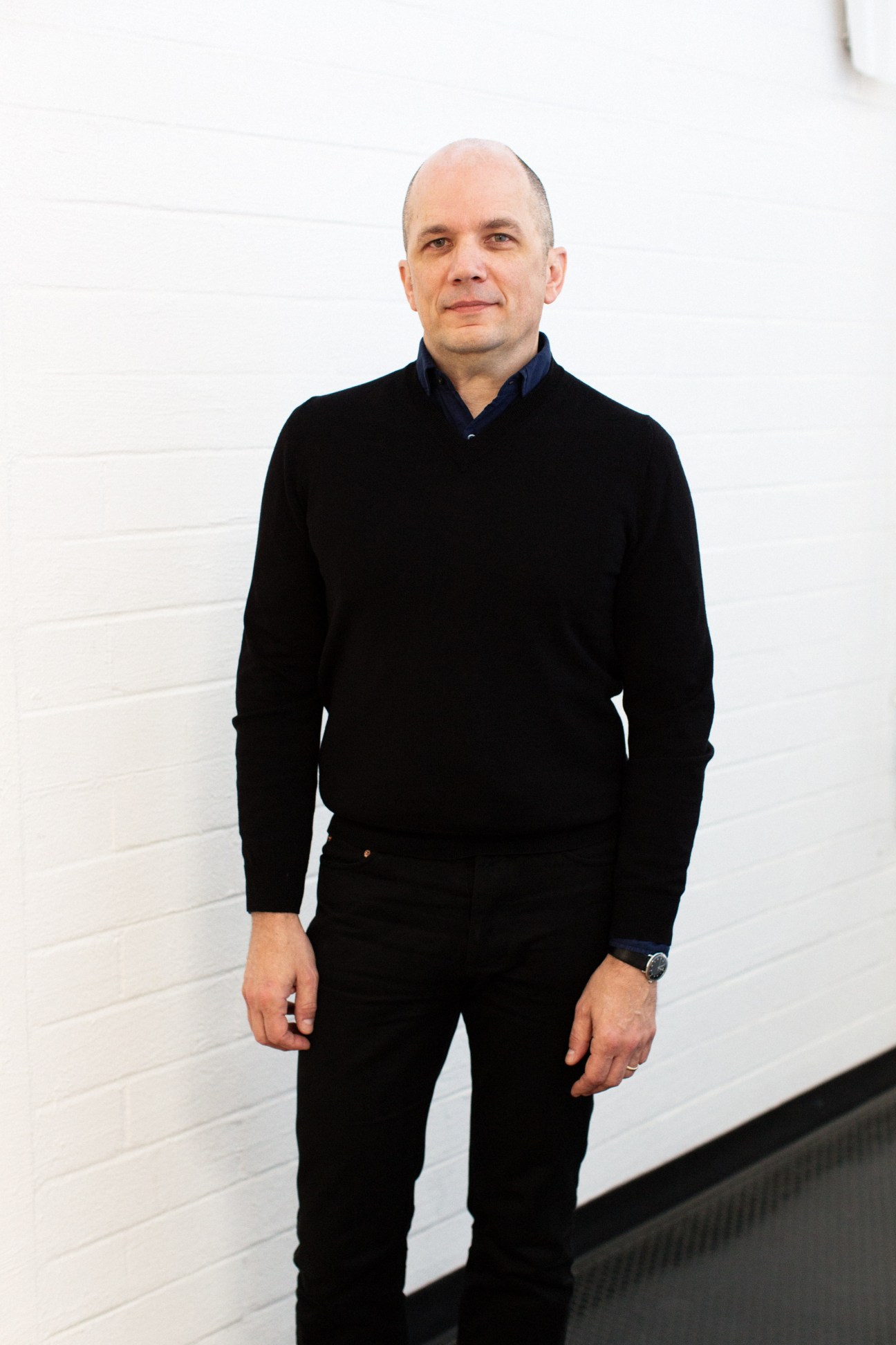 Portrait of architect Michael Maltzan standing against a white brick wall wearing black clothing.