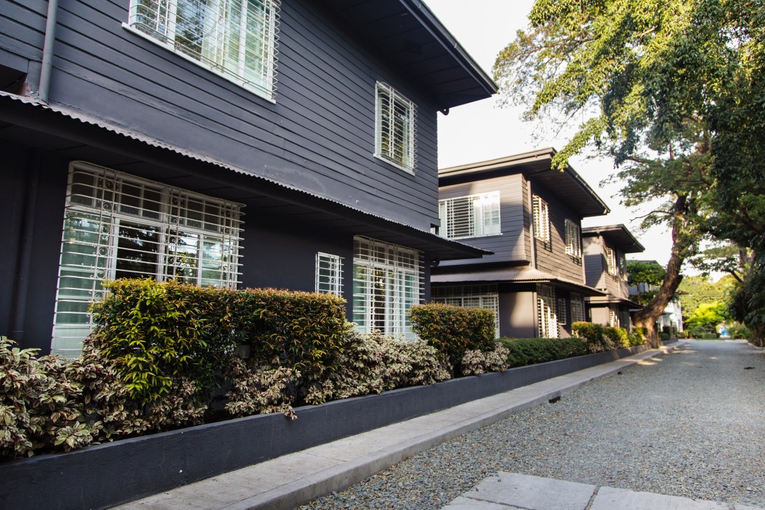 Modern residential buildings with dark exterior cladding, metal railings, and manicured hedges lining a paved pathway.