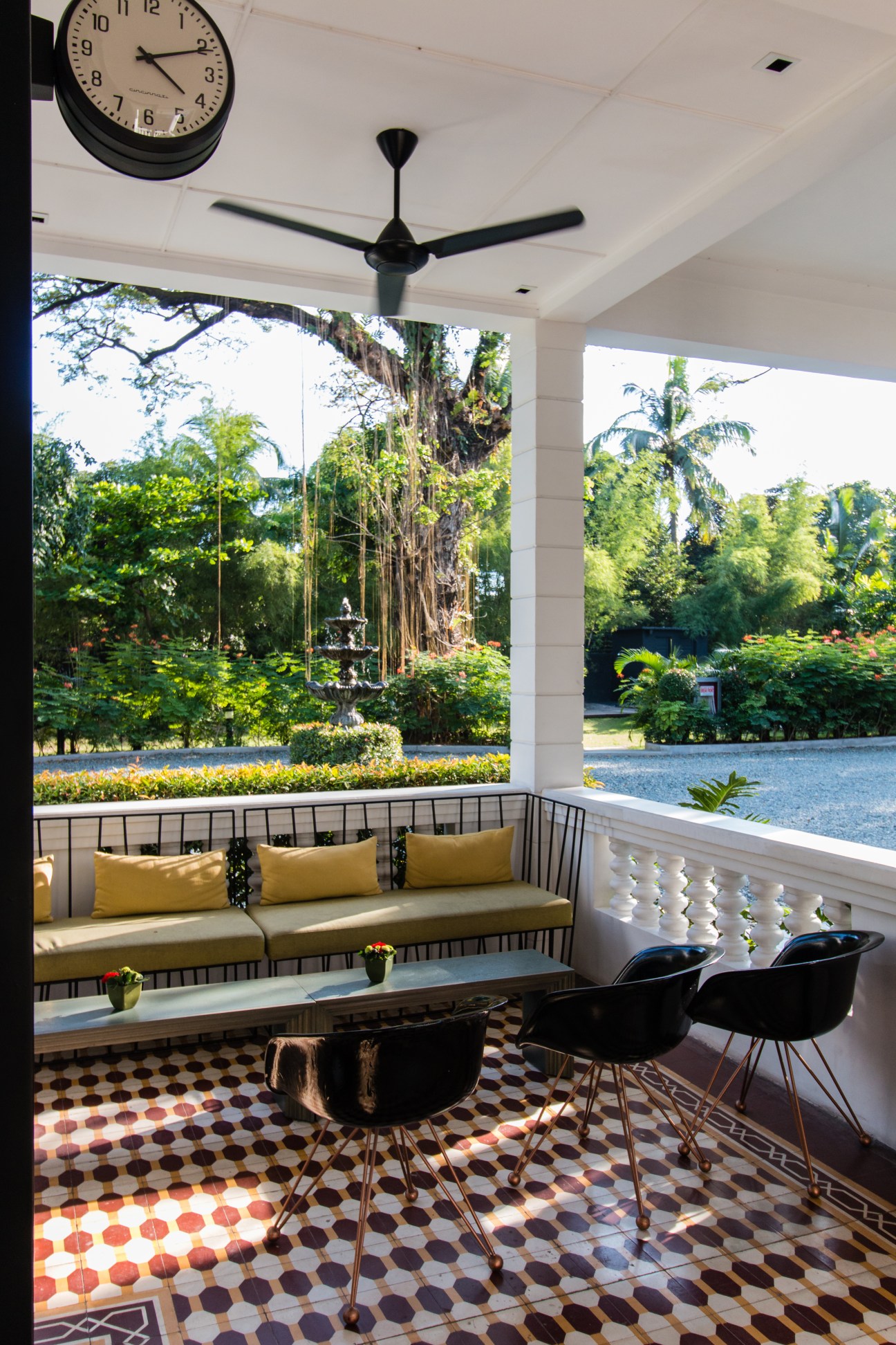 Modern covered porch with yellow cushioned seating, black chairs, geometric tile flooring, and views of tropical gardens.