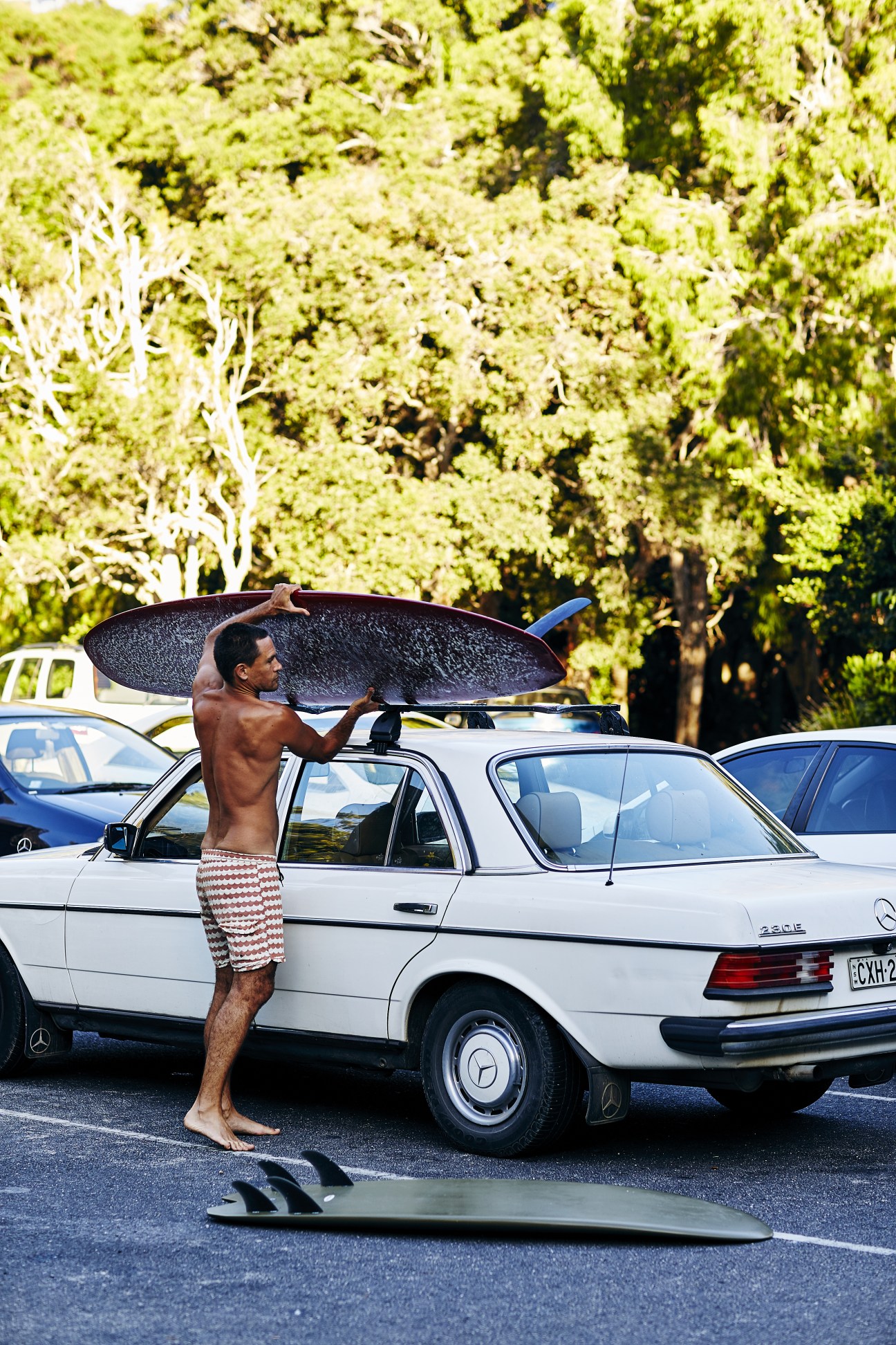 Shirtless surfer Marcello Sanó holds a black surfboard while standing next to a white Mercedes car in a parking lot with lush green forest in the background.