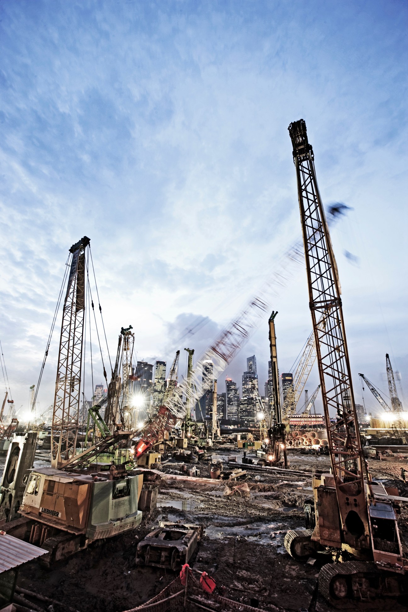 Construction site with tall piling cranes and excavation equipment with Singapore skyline in background.