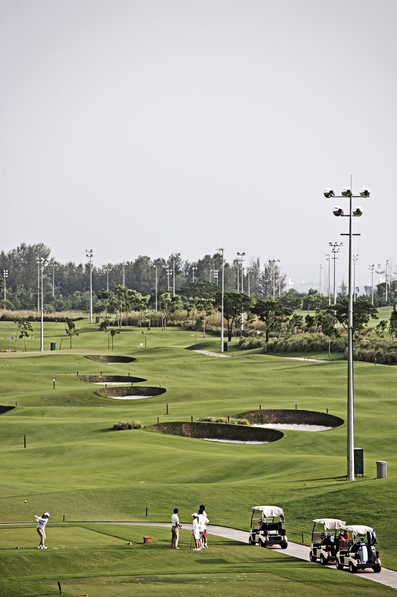 Golfers and carts on a manicured fairway with bunkers, trees and floodlights under overcast sky.