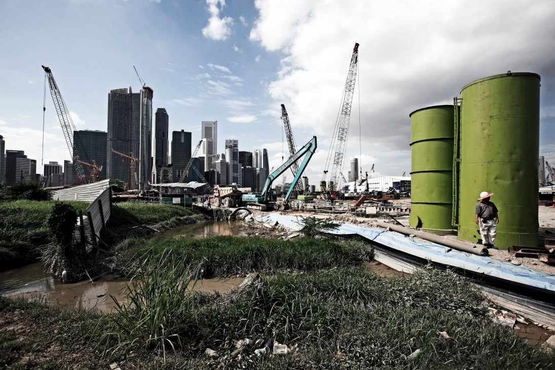 Construction site with cranes, green storage tanks, and Singapore's Marina Bay financial district skyline in the background.