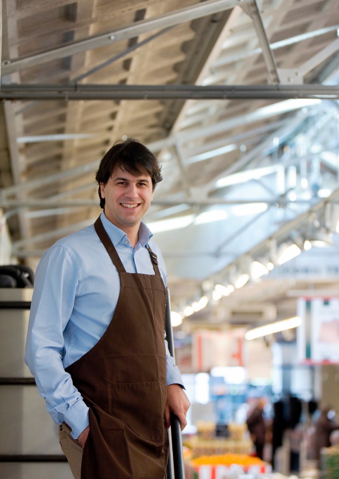 Man in blue shirt and brown apron smiling at camera in a market with glass ceiling.
