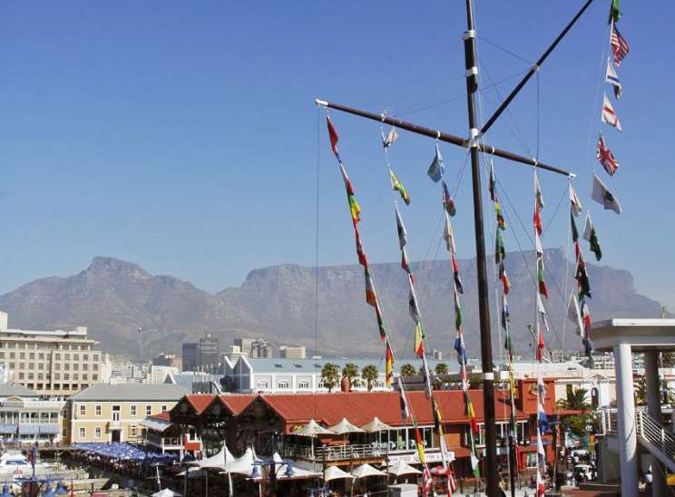 Waterfront promenade with international flags hanging from tall poles, mountains visible in background.