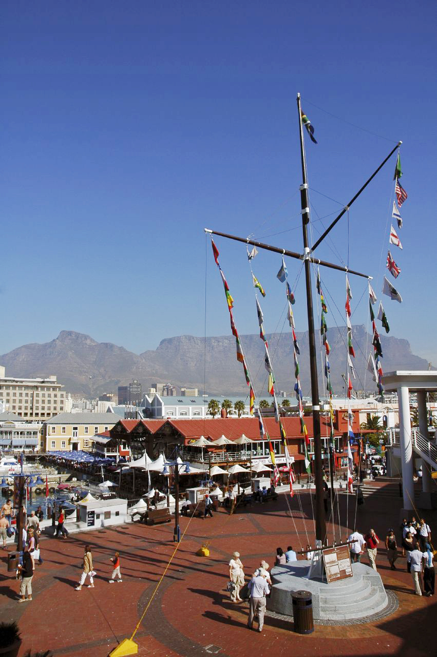 Waterfront promenade with international flags hanging from tall poles, mountains visible in background.