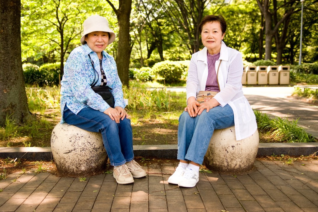 Two Japanese women in their mid-60s sitting on stone spheres in a park, smiling at the camera.