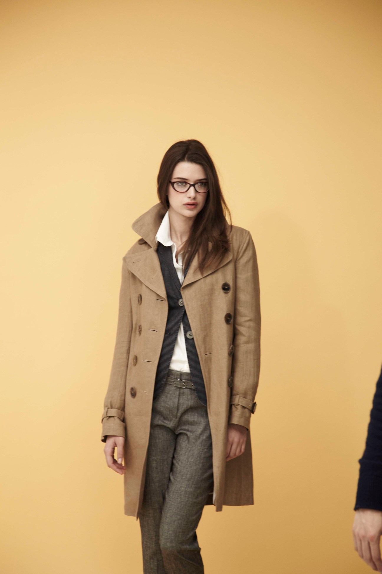 Woman wearing a beige double-breasted trench coat over a black waistcoat and white shirt, paired with grey trousers and glasses against a yellow background.