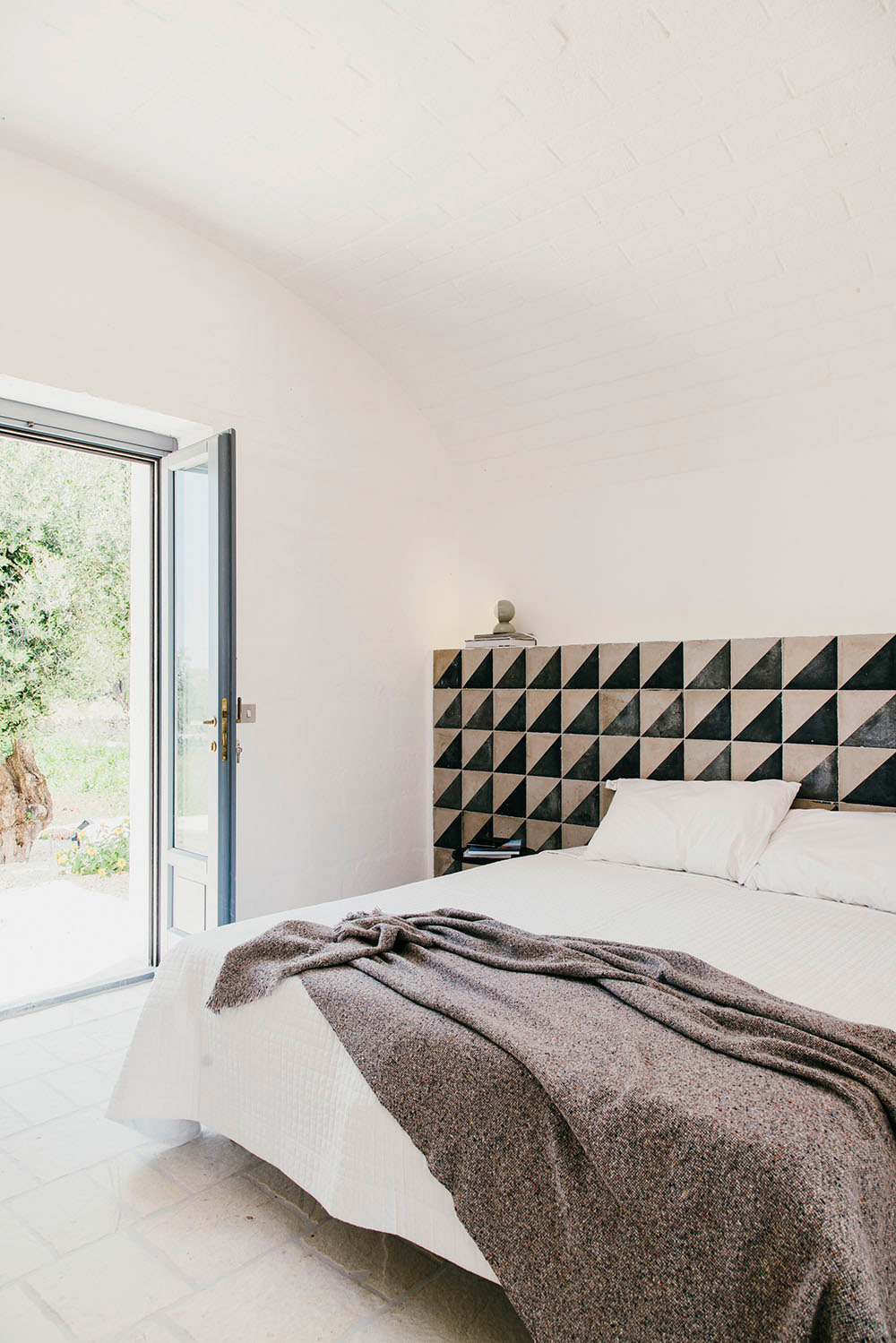 Minimalist bedroom with geometric patterned headboard, white bedding, brown throw blanket, and glass doors opening to greenery.