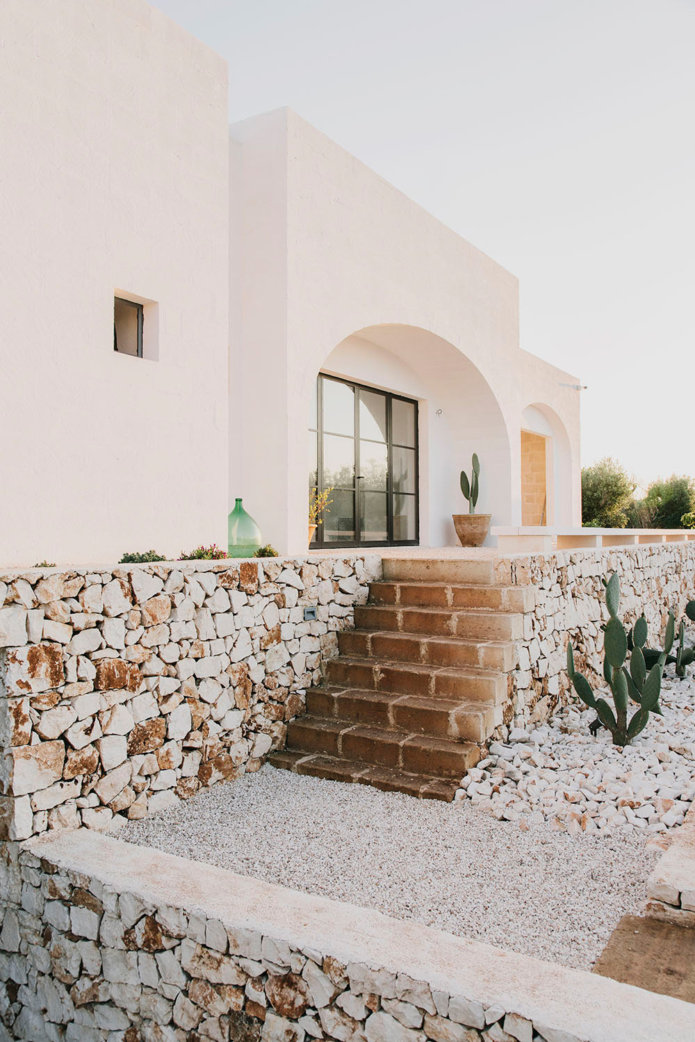 White-stone farmhouse with arched entrance, stone walls, and potted cactus in Puglia countryside.