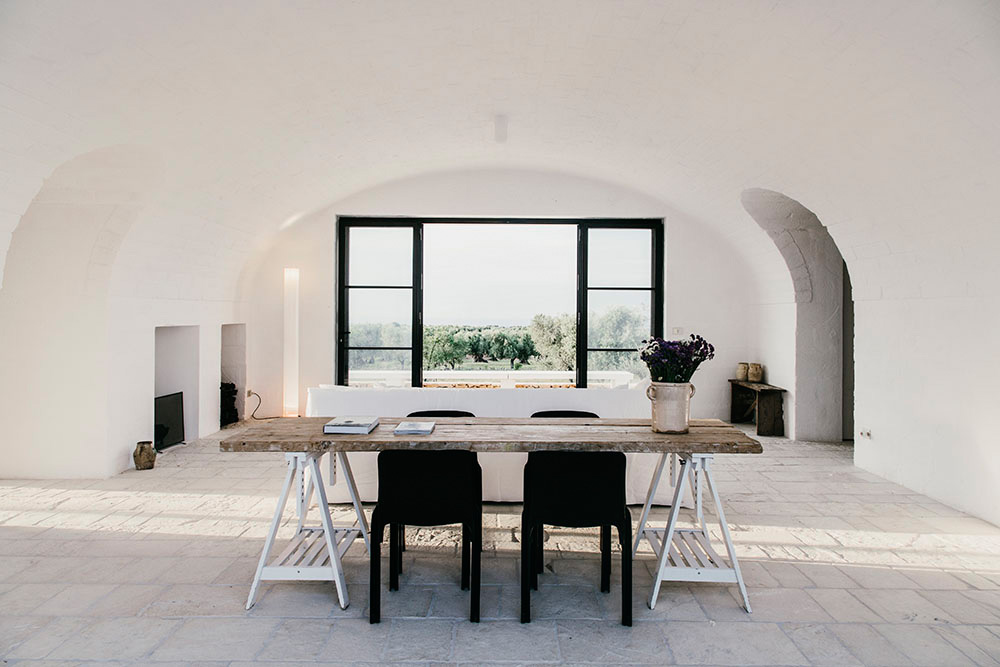 Minimalist interior with white stone walls, arched ceilings, wooden table, black chairs, and large windows overlooking olive groves.