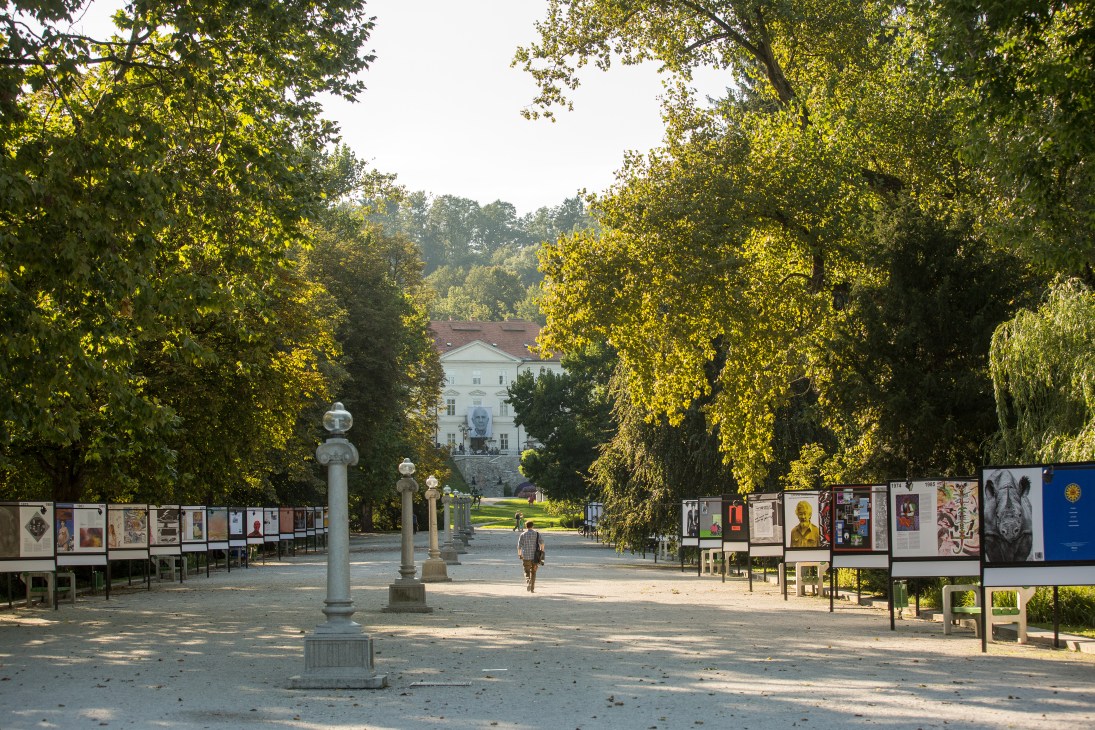 Posters displayed along a tree-lined promenade in Tivoli Park with classical building visible in background