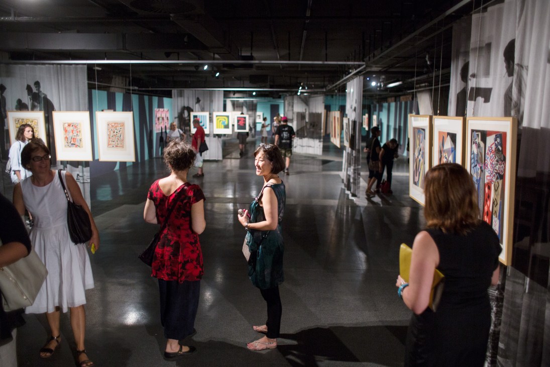 Visitors viewing framed artworks displayed on stands in a modern gallery space during Giles Round's exhibition.